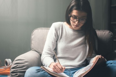 Κοπέλα με γυαλιά καθισμένη σε καναπέ και γυαλιά να σημειώνει σε τετράδιο